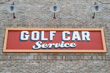 Fototapeta premium A red and yellow golf car service sign with white text and black trim. The sign is affixed to a brown and beige colored brick building with three barn lights over the signage for mechanical servicing.