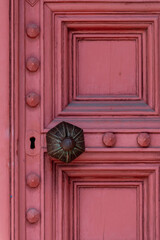 A maroon colored vintage wooden panel door with decorative ornate rosettes and molding. There's an antique metal tarnished door knob with patterns. The thick wooden door has a keyhole.