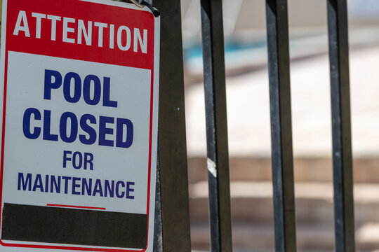 Attention pool closed for maintenance sign posted on a black metal fence with a swimming pool in the background. The warning indicates the pool is not available and a barricade is on the property. 