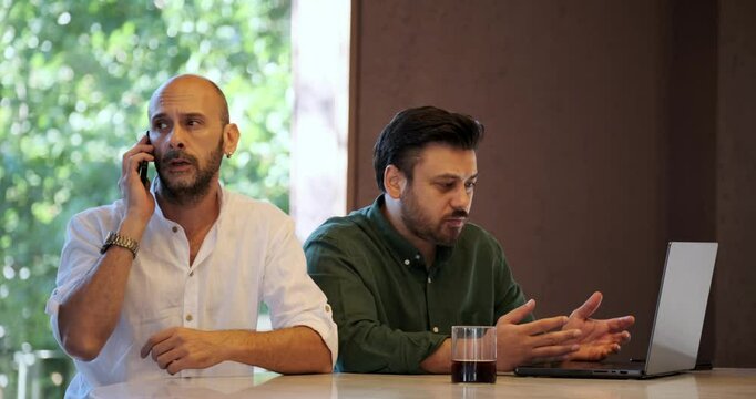 Two handsome middle-aged businessmen are having a meeting on their laptops outside the office environment. One of the men ignores the discussion and takes a phone call, which makes the other man angry