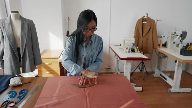 Professional seamstress working in fashion atelier, drawing patterns on fabric. Senior Asian woman creating exclusive handmade clothes, Chinese woman owner of tailor-made atelier in fashion business