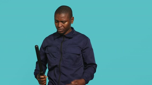 Portrait of security guard removing cane from belt while overseeing premises. Private law enforcement worker pointing to truncheon, threatening to subdue criminals, studio backdrop, camera A