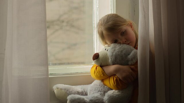 Sad little girl child sitting looking at window hugging teddy bear favorite toy childhood home unhappy kid daughter problem alone crisis pensive thinking mental health psychology stress upset suffer