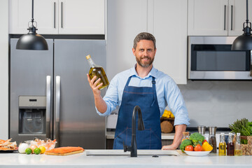 Handsome man in cook apron cooking with bottle olive oil in kitchen. Portrait of middle aged man in chef apron hold bottle olive oil in kitchen.