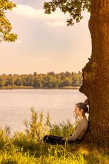 A woman relaxes near a lake under a tree on a sunny day, surrounded by natures beauty and tranquility