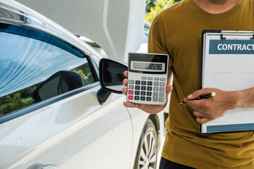 A businessman stands next to his damaged vehicle after an accident, holding a clipboard and...