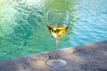 A glass of white wine stands on the edge of the swimming pool. Sun rays, light, shadows and blue water. Close-up