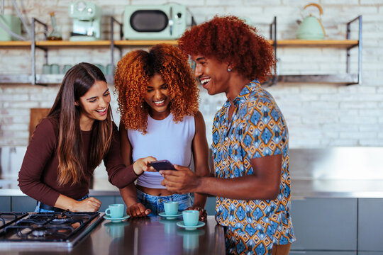 Three friends laughing at something on the phone inside of a kitchen