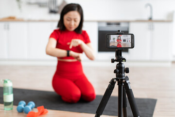 Sporty woman in red outfit talks about smart watch on camera. Filming fitness content in kitchen. Closeup of smartwatch with workout equipment in foreground.