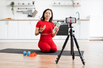 Woman in red activewear filming fitness vlog in kitchen. She explains exercise routines using weights, resistance bands, and water bottle for hydration. Bright, clean kitchen backdrop.