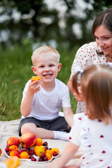 A boy with blond hair eats apricots and fruits, sitting next to a smiling mother and sister at a picnic. The concept of family recreation, health