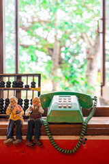 An antique green push button telephone is placed on a shelf with dolls of an uncle and aunt making a phone call with a green tree background in a blurred lens.