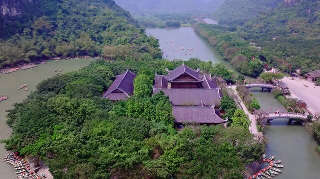 Serene aerial view of a temple surrounded by lush greenery in Tam Coc, Ninh Binh, Vietnam. The shot features traditional architecture and a peaceful river with boats.