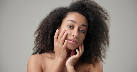 Beauty and healthcare concept - beautiful African American woman with curly afro hairstyle and clean, healthy skin touches her cheek and face with her hand, posing and looking at the camera