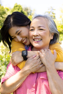 Asian woman hugging senior mother from behind, both smiling in outdoor setting