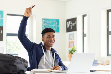 In school, male student raising hand while using laptop in classroom, smiling