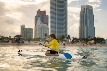 Kid riding on a paddle board in Miami Beach. Summer holidays. Kid paddle surf surfer little blonde...
