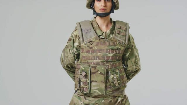 Camera tilts up to show young female soldier in uniform wearing body armour and helmet in front of plain studio background - shot in slow motion