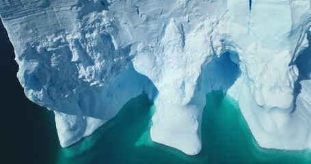 Fototapeta premium Giant ice wall melting in arctic water aerial shot. Antarctica iceberg floating turquoise water top down view. Huge glacier ice cave melting in the ocean close up. Global warming and climate change.