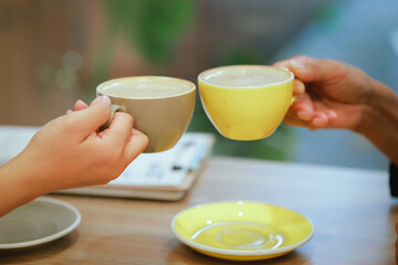 Asian man and woman, enjoy a morning meeting at a cafe, holding cups of hot lattes, clinking together in a cheerful, relaxed atmosphere with a blurred background.