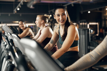 Fit young woman in sportswear laughing while riding on a stationary bike with a group of smiling female friends during a cardio workout session in a gym