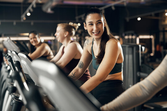 Fit young woman in sportswear laughing while riding on a stationary bike with a group of smiling female friends during a cardio workout session in a gym