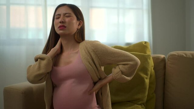 Young pregnant woman suffering from backache and shoulder pain alone at home, asian female touching her back during pregnancy, accidental medical urgency case and health care insurance for pregnancy.
