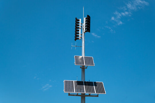 Poste com uma antena, m&uacute;ltiplos alto-falantes e pain&eacute;is solares para transmiss&atilde;o de alertas sonoros com o c&eacute;u azul como fundo