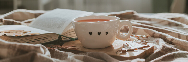 Cozy autumn at home. Glass cup with hot tea, open book and autumn leaves on blanket on bed. Long banner for design, website