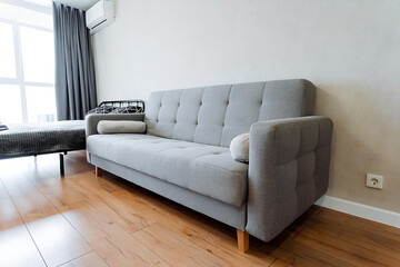 A grey couch is positioned in a living room next to a window, amidst subtle elements of furniture, building features, curtains, and wood providing a cozy and inviting atmosphere