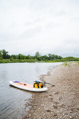 A paddle board rests on the riverbank under the open sky. It represents tranquility and adventure, a perfect blend of nature and watercraft, providing a serene scene by the water