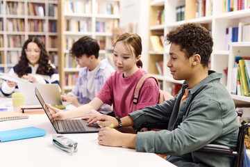 Side view portrait of smiling young boy using laptop in school library with diverse group of students copy space