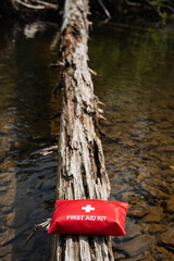 A red first aid kit is positioned on a weathered log in a tranquil creek, surrounded by nature and wilderness. It evokes a sense of serenity and safety in the remote outdoors