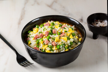 Cantonese Fried Rice and egg omelette with green pea, ham steak, spring onion, ribe long rice in a beige bowl on a white marble table, served with fork. Eurasian cuisine.