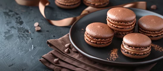 Gordijnen Dark chocolate macarons displayed elegantly on a black plate with a ribbon and a brown napkin, set on a dark table background with copy space image. © HN Works