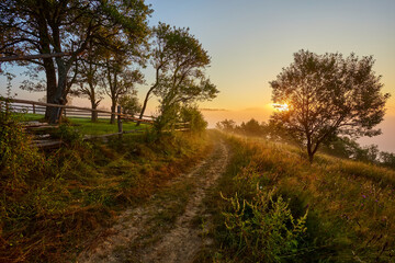 Beautiful dreamy autumn sunrise rural scenery. Haystacks and trees on a mountain hill with fog.