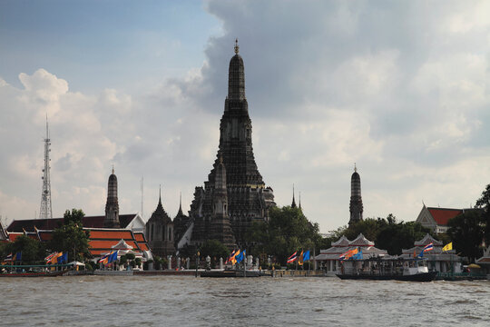 View of Wat Arun Ratchawararam Buddhist temple by the river with boats and flags, Bangkok, Thailand.