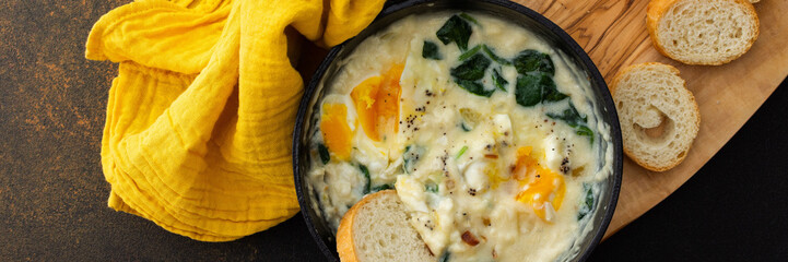 Healthy breakfasts or dinners banner, fried eggs with cheese, spinach leaves and coconut milk in a cast-iron pan, sliced baguette on a cutting board on a brown background