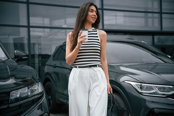 With white cup of drink. Attractive young woman is outdoors near her brand new car