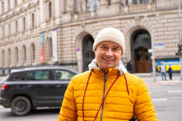 Cheerful middle-aged man, 65 in winter bright yellow puffer jacket and beanie, smiling on urban...