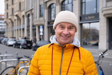 Cheerful middle-aged man, 65 in winter bright yellow puffer jacket and beanie, smiling on urban street with historic buildings in background © kittyfly
