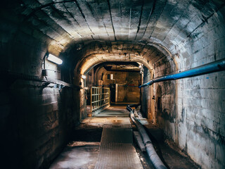 Obraz premium Industrial underground tunnel with metal walkway and pipes, illuminated by dim artificial light.