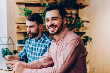 Portrait of cheerful hipster guy looking at camera studying in coworking space with colleague using laptop computer, prosperous young entrepreneurs earning via internet on freelance together