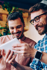Cheerful young hipster guys having fun watching funny videos from social networks via touchpad together, smiling bearded man showing his results to friend playing online games on portable pc