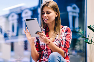 Young woman reading information from tablet working on completing project before deadline in office, hipster girl sitting on window sill with portable pc watching webinar preparing for exams overtime