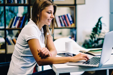 Smiling hipster girl satisfied with useful online language courses watching videos for learning via netbook in college library, positive female student using laptop computer for checking latest news