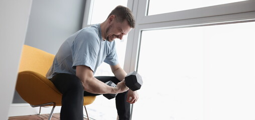 Young man sitting on chair and lifting heavy dumbbell. Morning home sports workouts concept