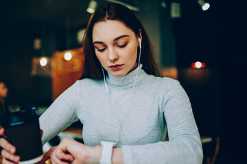 Pensive teen girl looking at touch screen of smartwatch checking notification about message,female using modern gadgets on coffee break listening music in earphones and managing time on digital clock