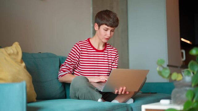 Busy Caucasian freelancer sitting in lotus position on sofa in living room. Hardworking woman holding laptop on legs while working online. Looking with concertation at display. Remote job concept.