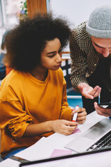 Bearded designer showing design application installed on smartphone to africa american colleague sitting at laptop computer in office for productive work of developing creative project
